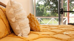 Ein Bett mit einer gelben Tagesdecke und zwei Kissen, eines weiß und eines mit Ananas-Muster, in einem Zimmer mit Blick auf Grün durch eine Glastür.