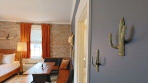 Een kamer met een bed, een bureau en een stoel. Er is een raam met gordijnen, een lamp en een cactusdecoratie aan de muur.
