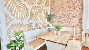 Comedor con mesa de madera y banco blanco, pared de ladrillo y plantas en macetas.