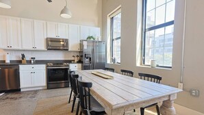 A modern kitchen with white cabinets, stainless steel appliances, a wooden dining table, and large windows. - Urban Retreat w/ Downtown Views (Knoxville)