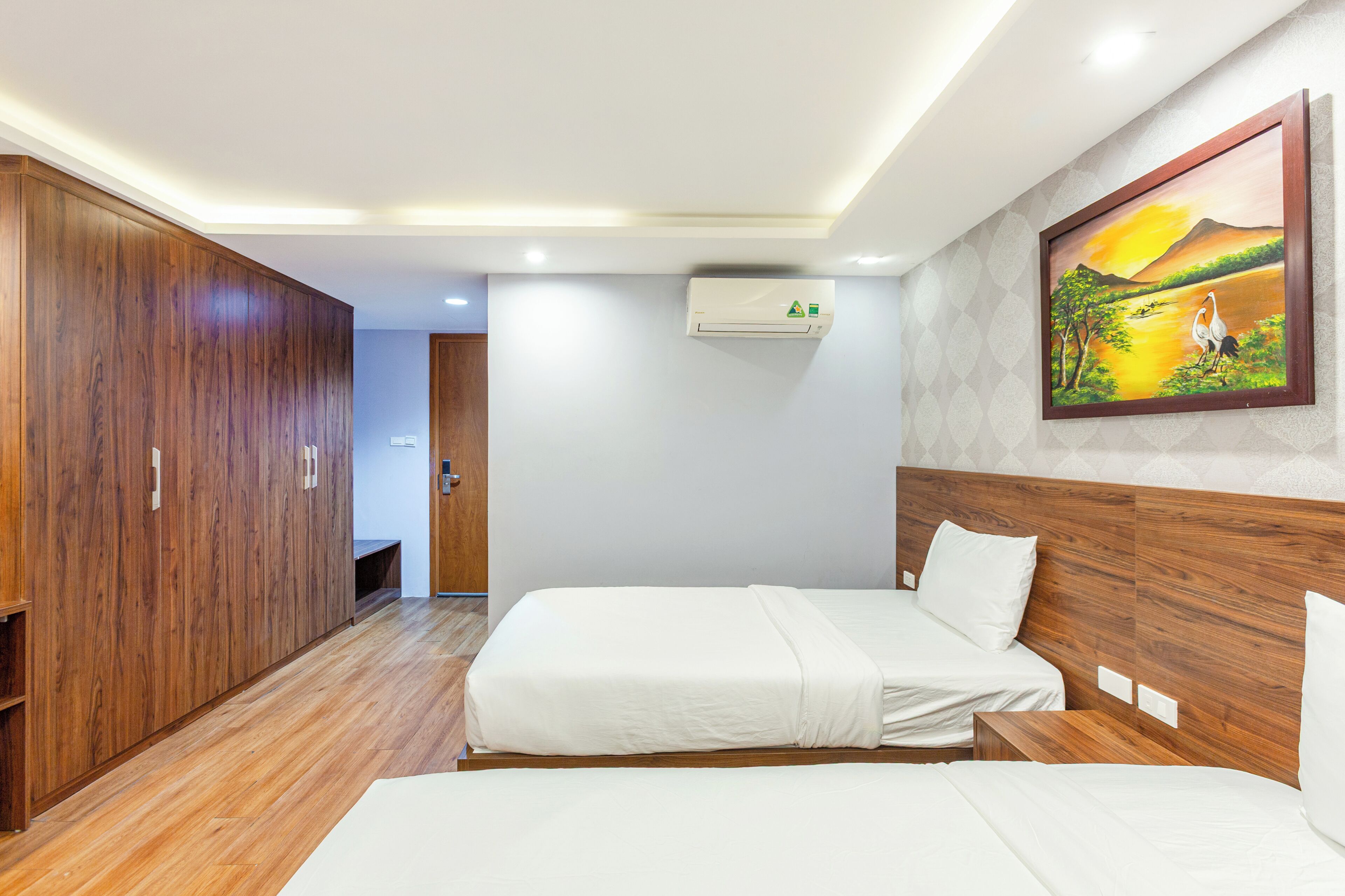 A hotel room with two beds, wooden wardrobe, an air conditioning unit, and a painting on the wall.