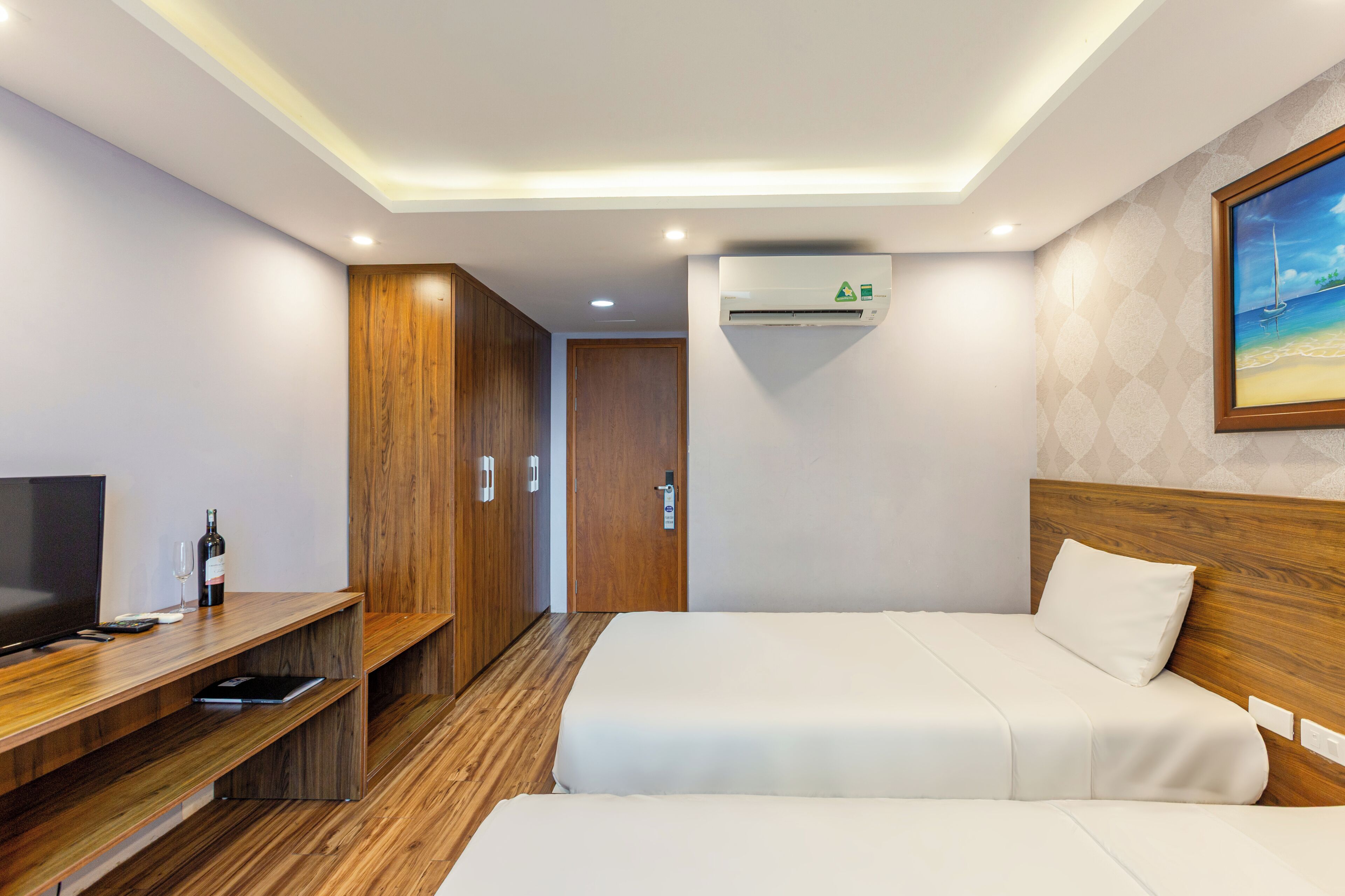 A hotel room with two beds, a TV, a desk with a bottle and glasses, wooden wardrobe, and an air conditioning unit.