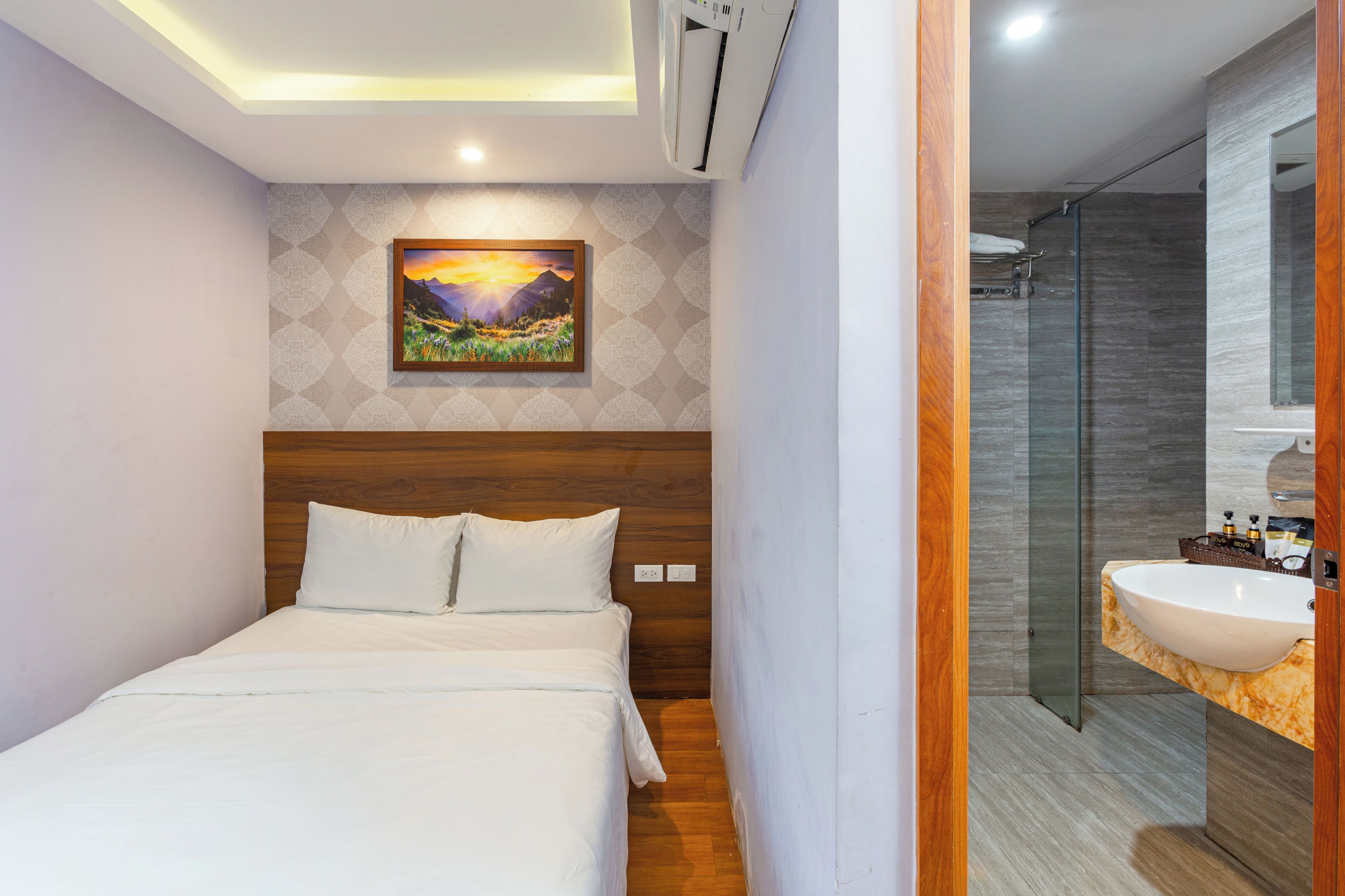 A hotel room with a bed, a painting on the wall, a shower area, and a bathroom with a sink.