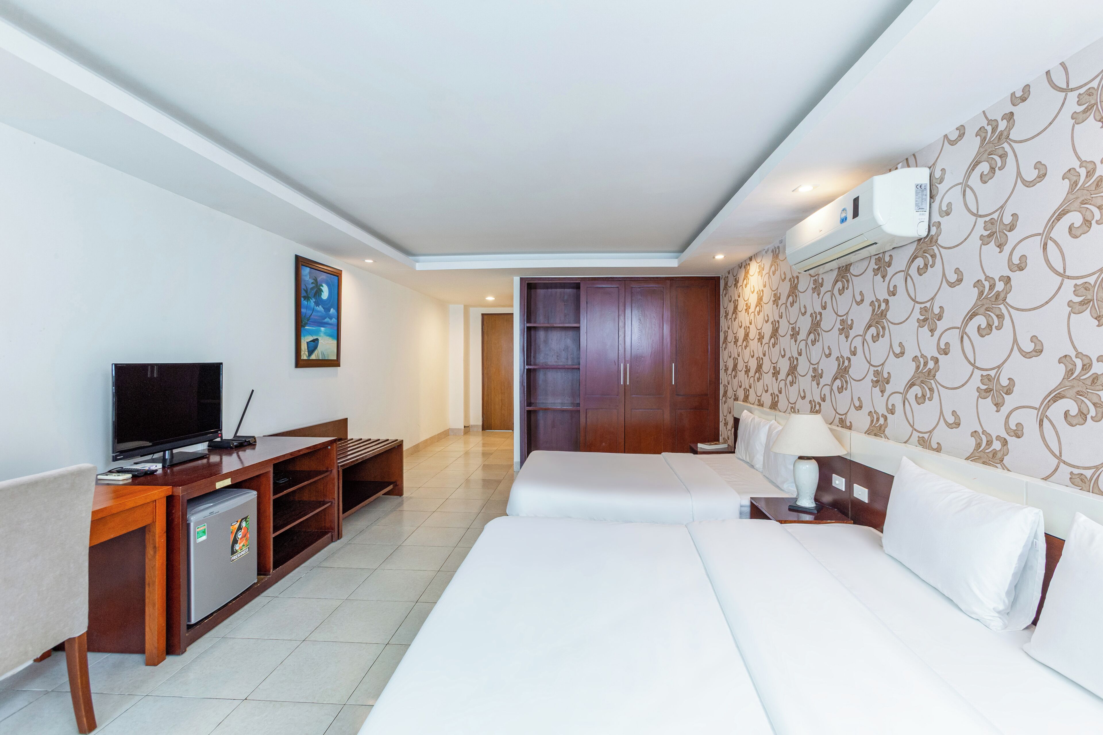 A hotel room with a bed, a desk with a television, a mini-fridge, and a wardrobe.