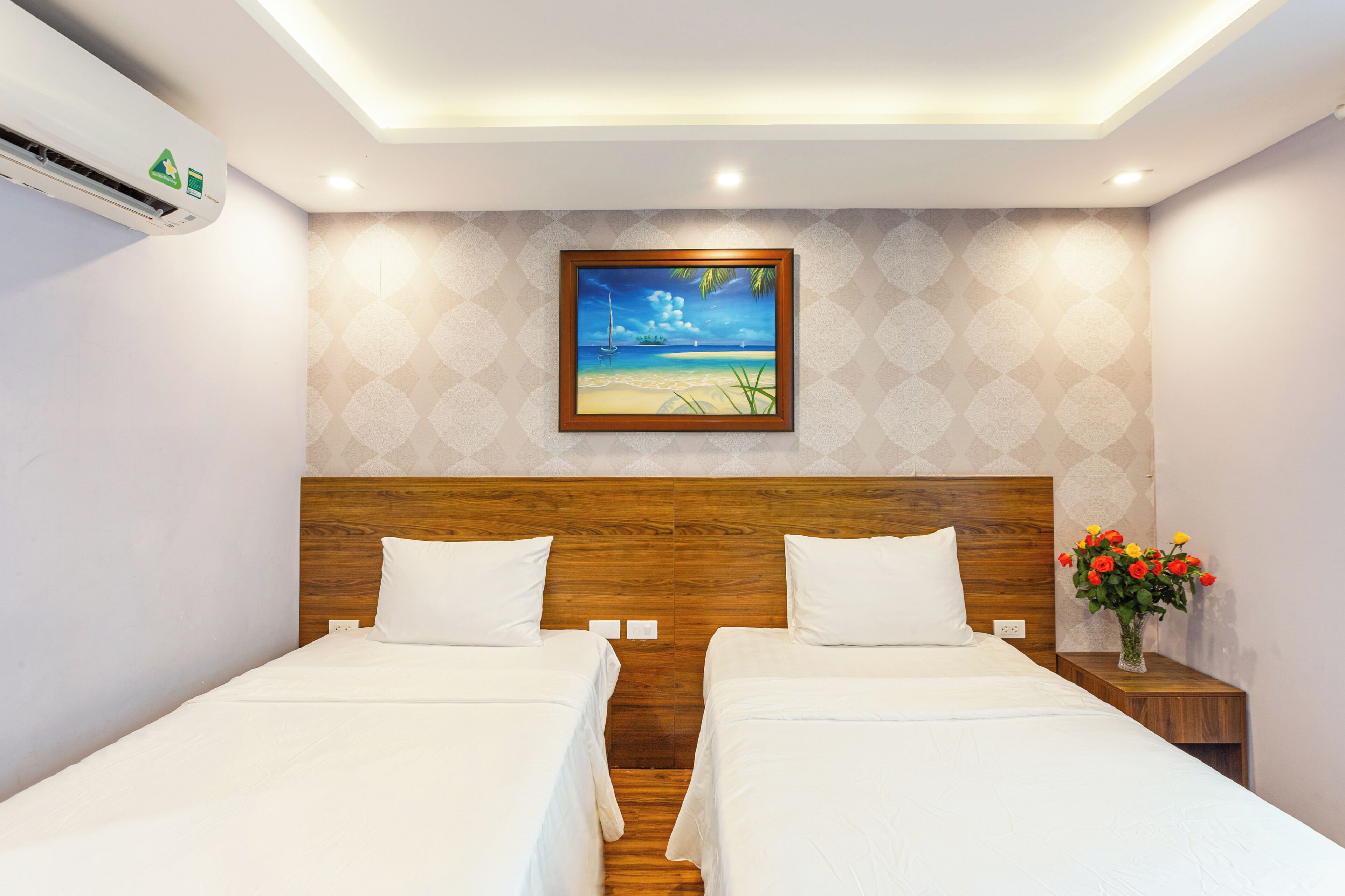 A hotel room with two beds, a painting of a beach scene, a nightstand with a vase of flowers, and an air conditioning unit.
