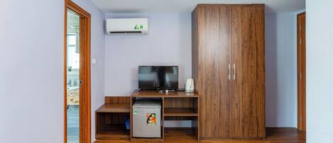 A room with a wooden wardrobe, a television, a small refrigerator, and an air conditioning unit on the wall.