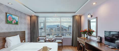 Hotelkamer met een bed, een bureau met een televisie, een zithoek en een groot raam met uitzicht op de stad.