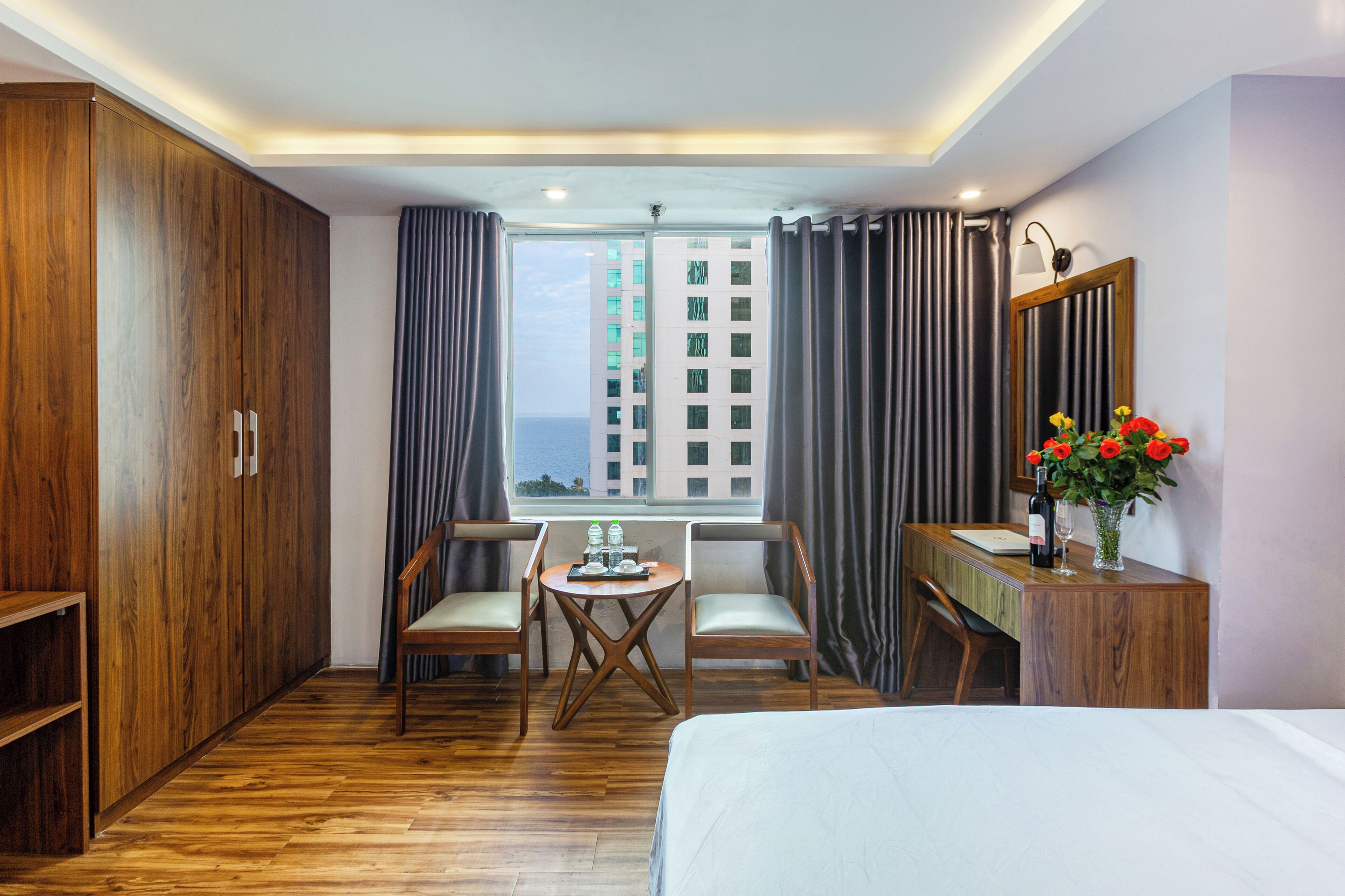 A hotel room with a wooden floor, a bed, two chairs, a small table with a bottle and glasses, a mirror, and a window with curtains.