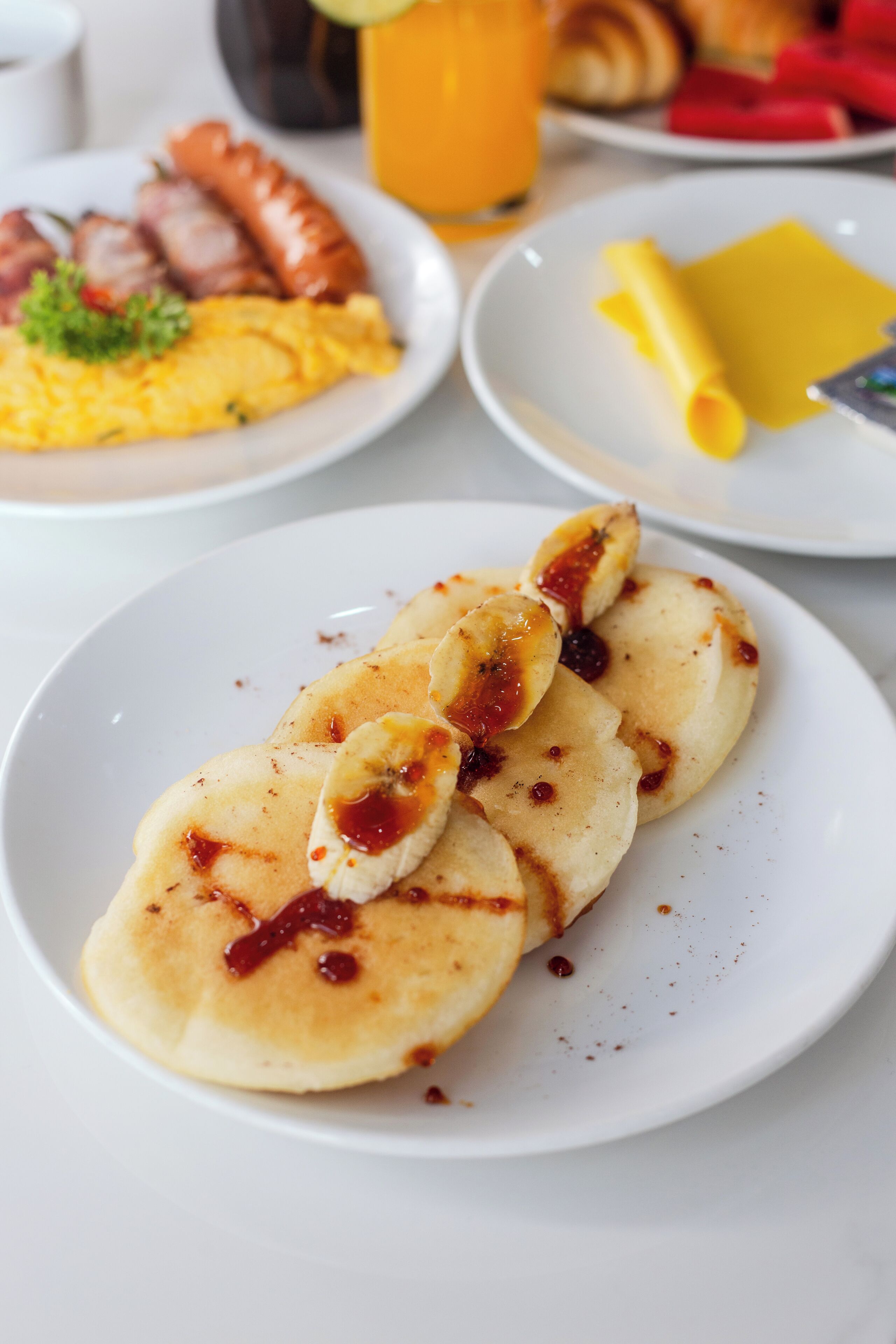 A plate of pancakes with banana slices and jam, a plate of sausages and scrambled eggs, and a glass of orange juice.