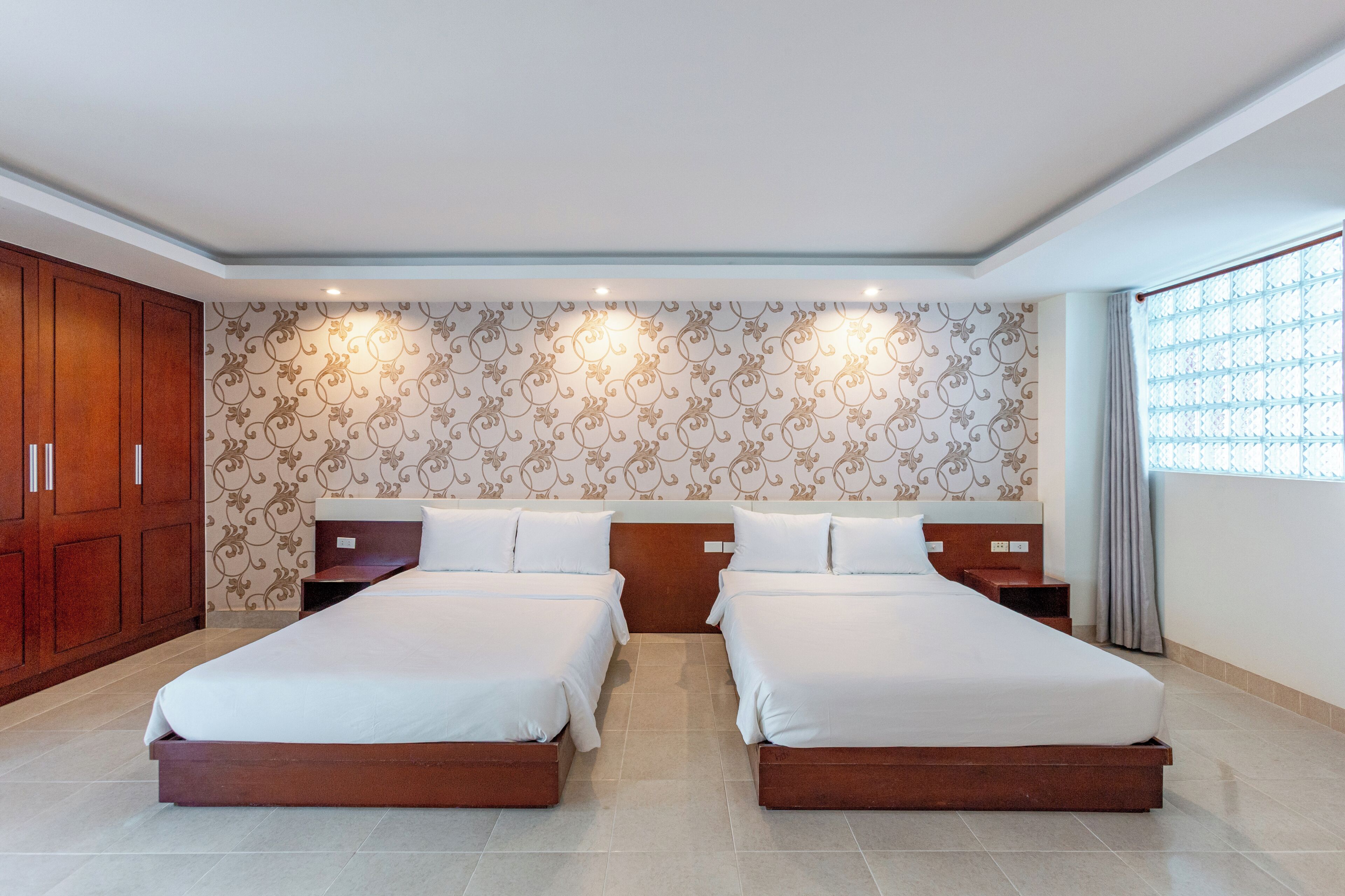 A hotel room with two beds, wooden wardrobe, patterned wallpaper, and a large window with curtains.