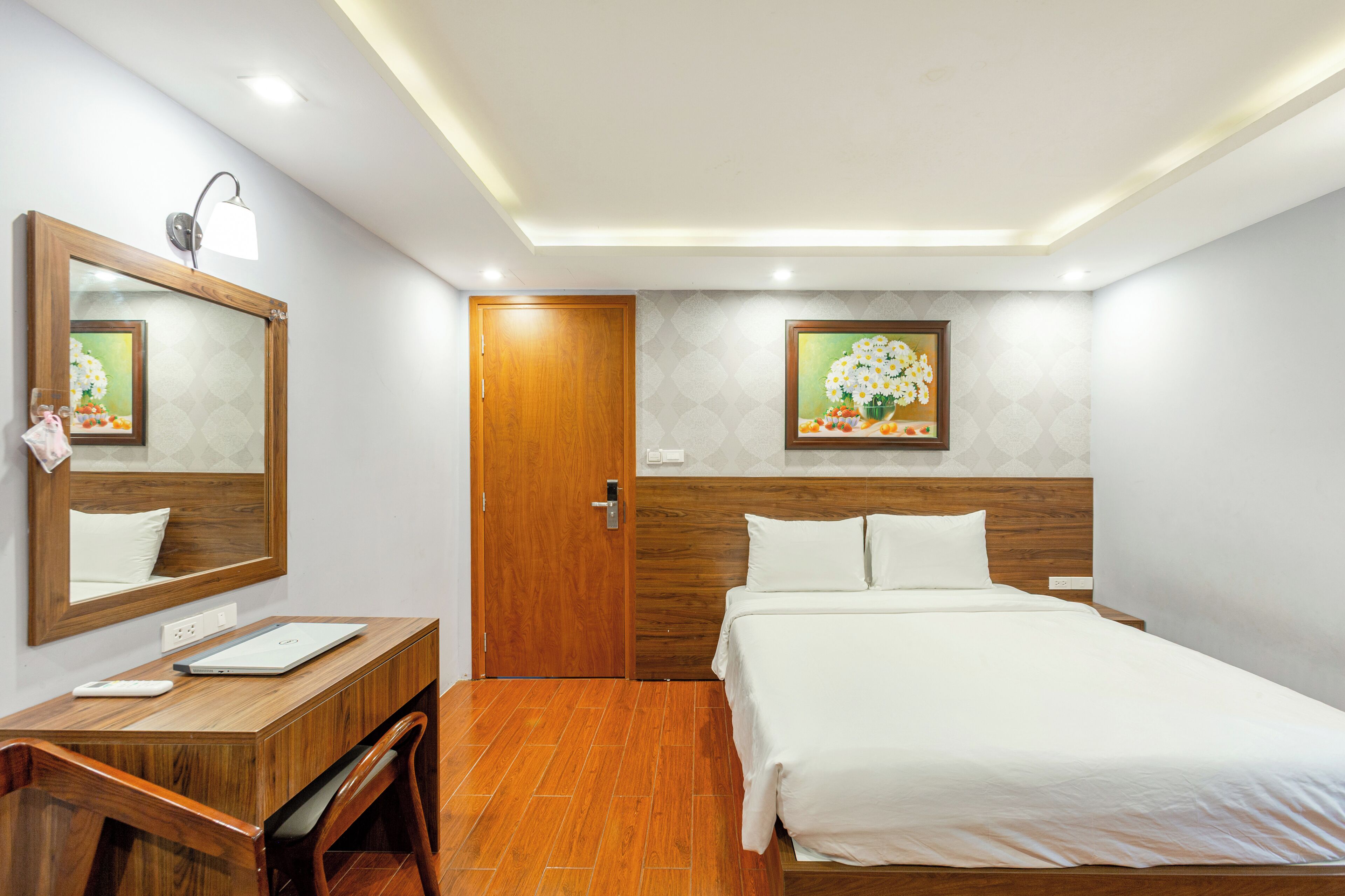 A hotel room with a wooden desk, a bed with white linens, a wooden door, and two wall-mounted paintings.