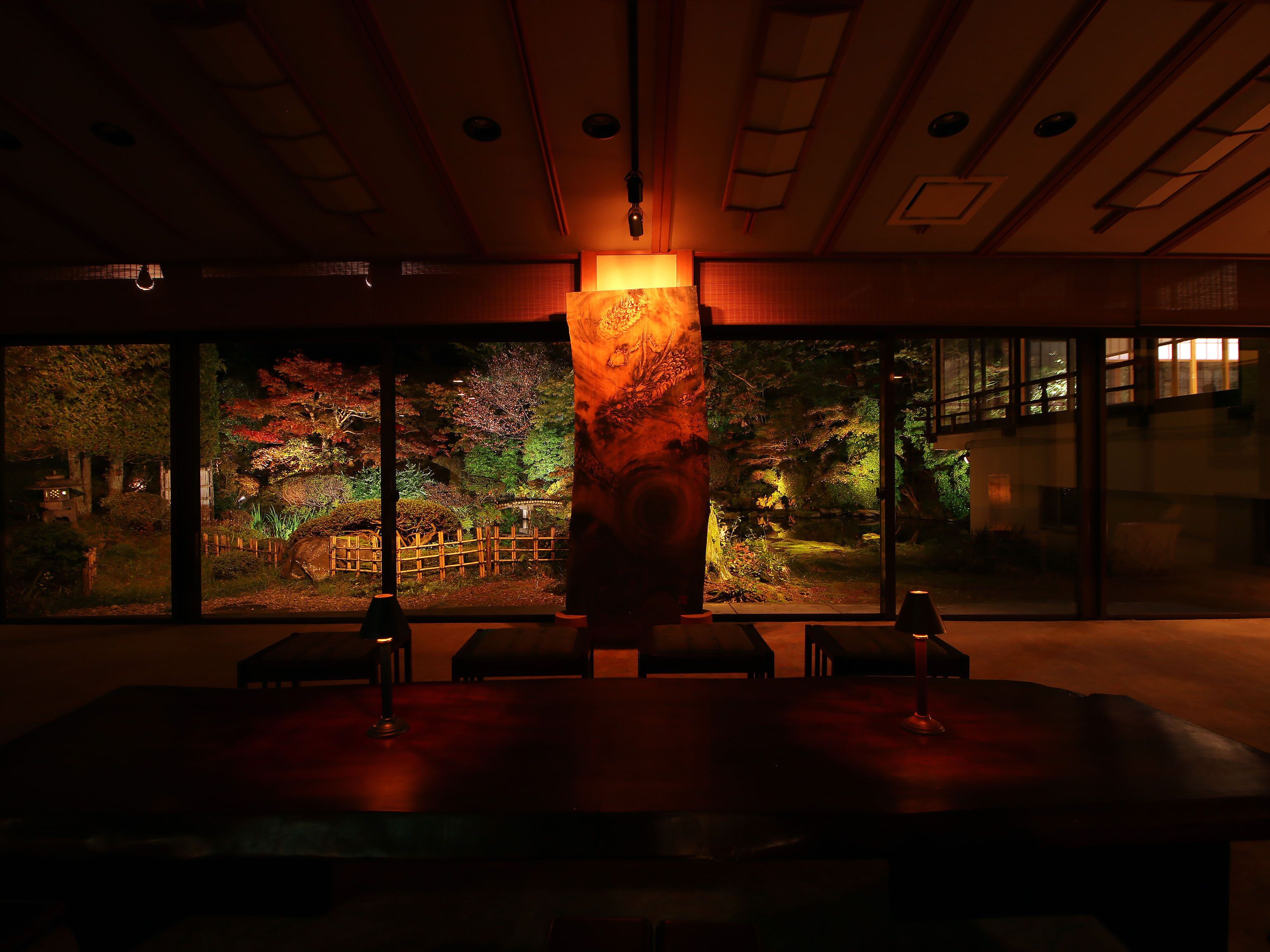 Una habitación con poca luz, un ventanal grande, una mesa de madera y un pilar con detalles tallados.