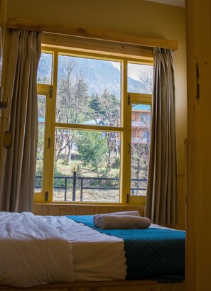 Una habitación con una cama, una ventana con cortinas y vistas a la montaña y árboles.