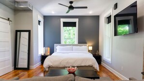Un dormitorio con cama, mesitas de noche, ventilador de techo, ventana y televisor montado en la pared.