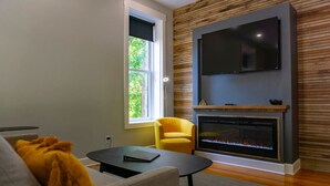 Una sala de estar moderna con chimenea, un televisor de pantalla plana montado sobre ella, un sofá, un sillón amarillo y un ventanal con vistas a la vegetación.