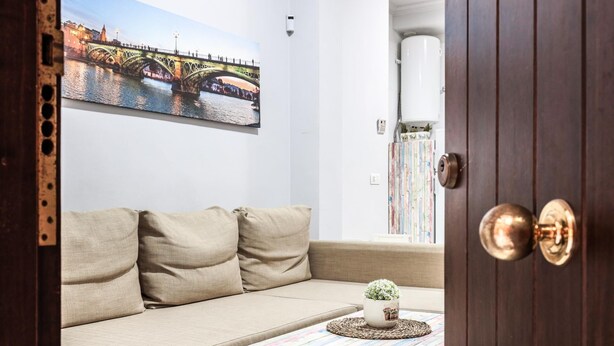 Um quarto aconchegante com um sofá bege, uma mesinha com um objeto decorativo e uma pintura de uma ponte na parede.