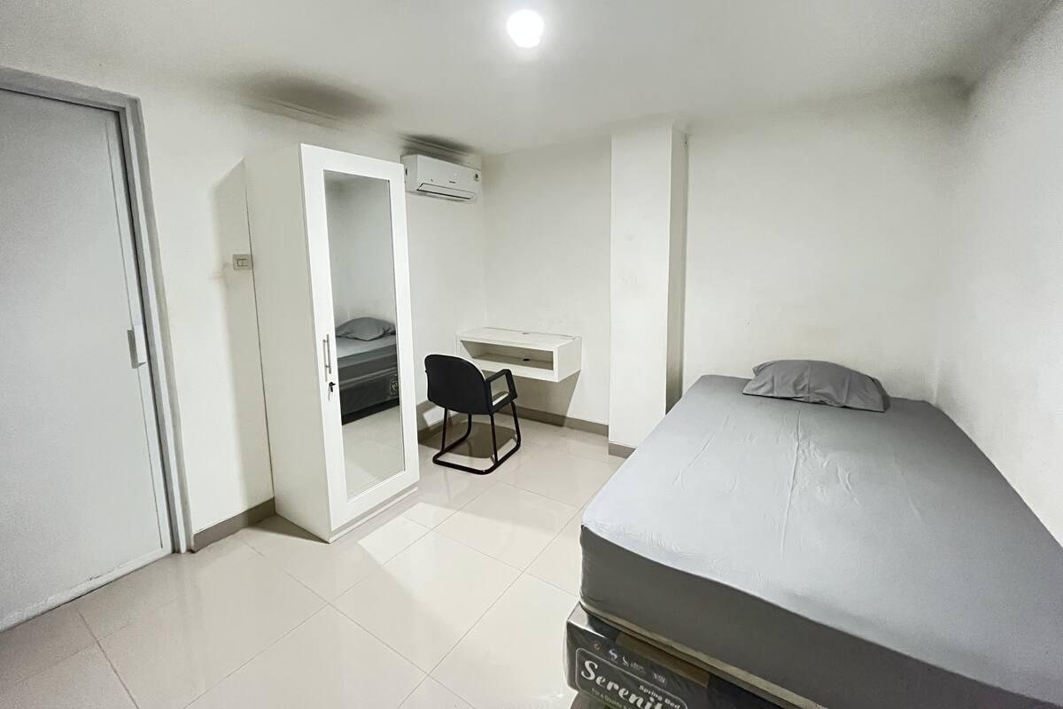 Un dormitorio con una cama, un escritorio y una silla. Se ve una puerta y un equipo de aire acondicionado en la pared.