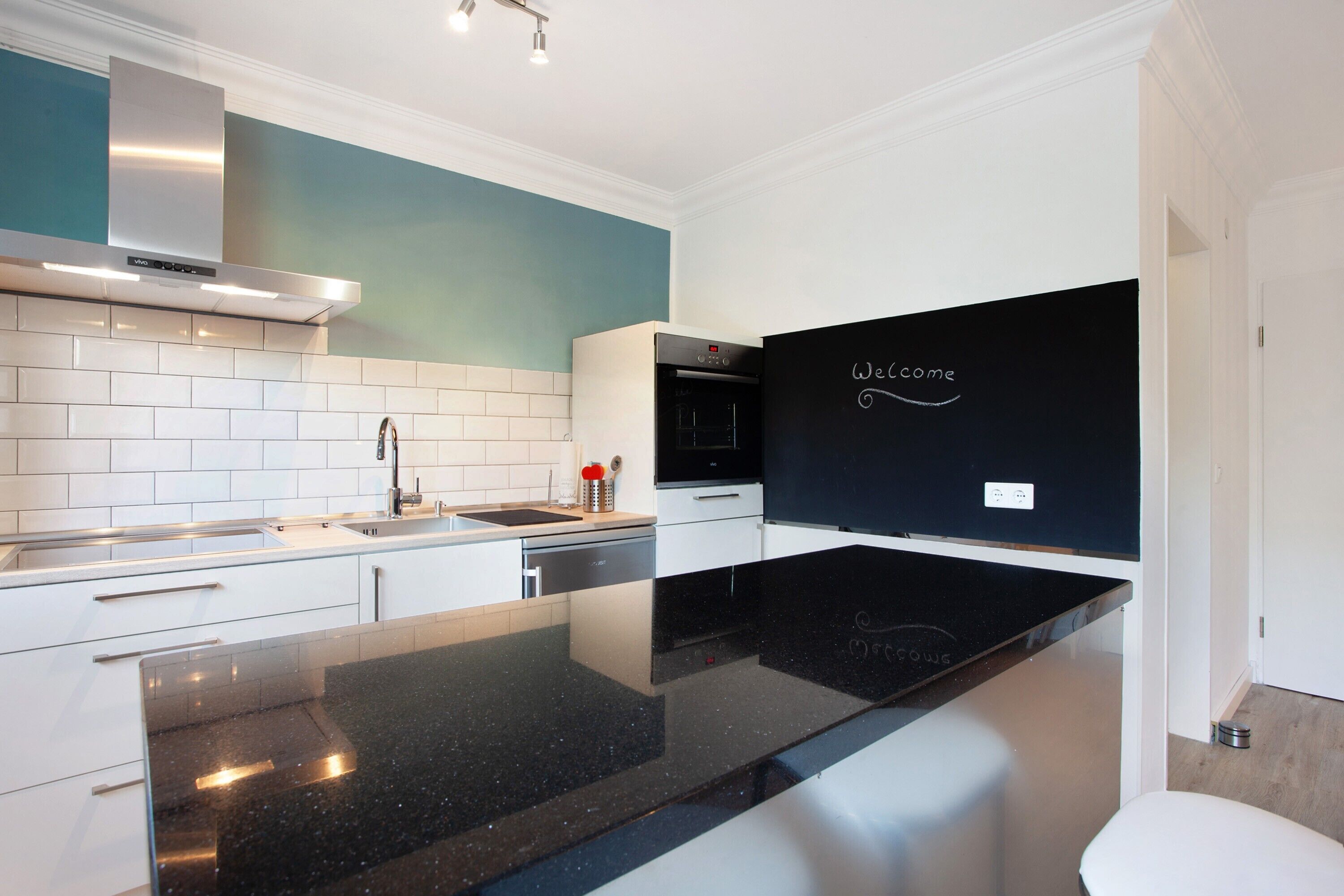 Een moderne keuken met witte kasten, een zwart aanrecht en een 'Welcome'-bord aan de muur.