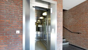 Un ascensor con puerta de cristal, pared de ladrillo y barandilla de metal.