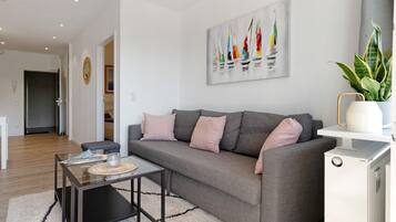 Un salon moderne avec un canapé gris, une table basse en verre, un radiateur blanc et un tableau représentant des voiliers accroché au mur.