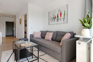 A modern living room with a grey sofa, a glass coffee table, a white radiator, and a painting of sailboats on the wall. - Apartment 'Ferienappartement Holm Ostsee' with Balcony and Wi-Fi (Schönberg-Holm)