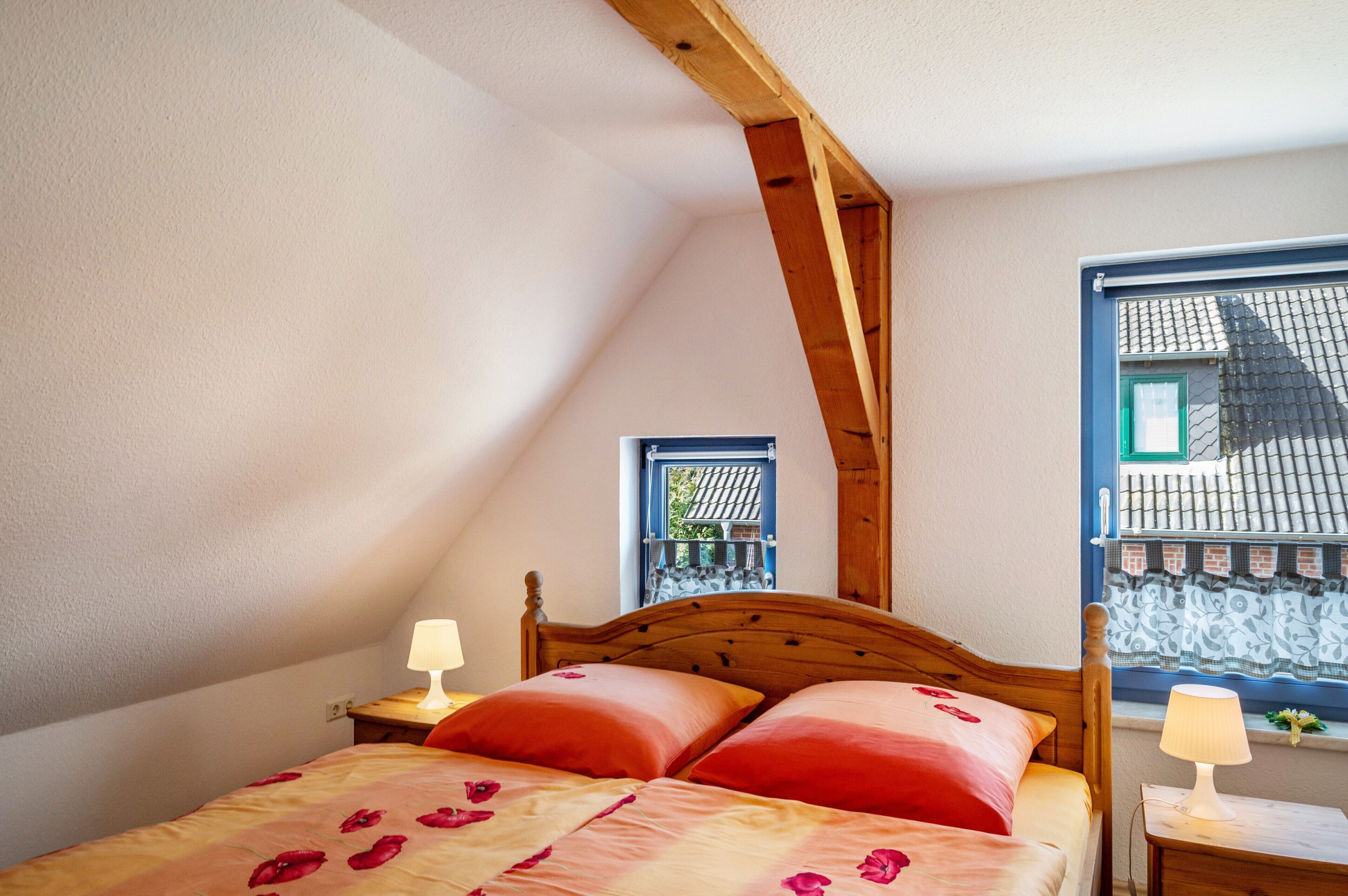 Ein Schlafzimmer mit schrägen Wänden, einem Holzbettgestell, zwei Nachttischlampen und einem Fenster mit Blick auf ein Gebäude.