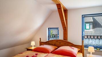 Ein Schlafzimmer mit schrägen Wänden, einem Holzbettgestell, zwei Nachttischlampen und einem Fenster mit Blick auf ein Gebäude.