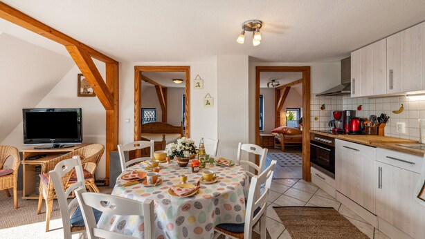 A dining area with a table set for six, a television, and a kitchen with white cabinets and a wooden countertop. - Apartment "Zur Eule - Uhu" with Shared Garden & Wi-Fi (Satow OT Clausdorf)