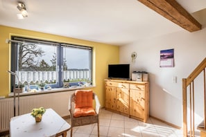 A room with a large window, a television, a wooden cabinet, a wicker chair, and a small table with a plant.