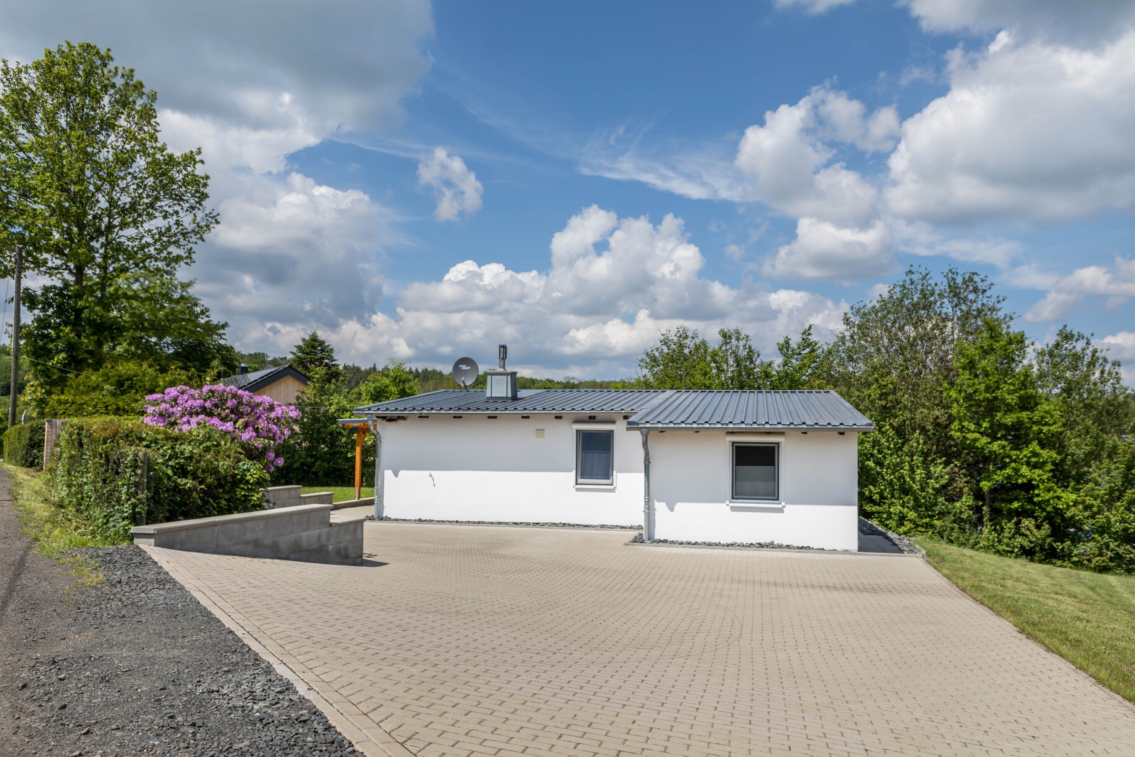 A small, single-story building with a paved driveway and a garden area with flowering plants.