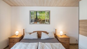 Un dormitorio con una cama, dos mesitas de noche con lámparas, un cuadro colgado en la pared que representa un paisaje forestal y un techo de madera.