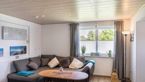 Una sala de estar moderna con techo de madera, un sofá gris con cojines, una mesa de centro de madera, una lámpara de pie y una ventana con cortinas.