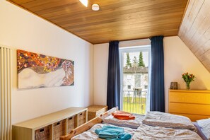 Un dormitorio con techo de madera, una cama con manta estampada, una mesita de noche, un cuadro colgado en la pared y vistas a un edificio y árboles a través de una ventana.