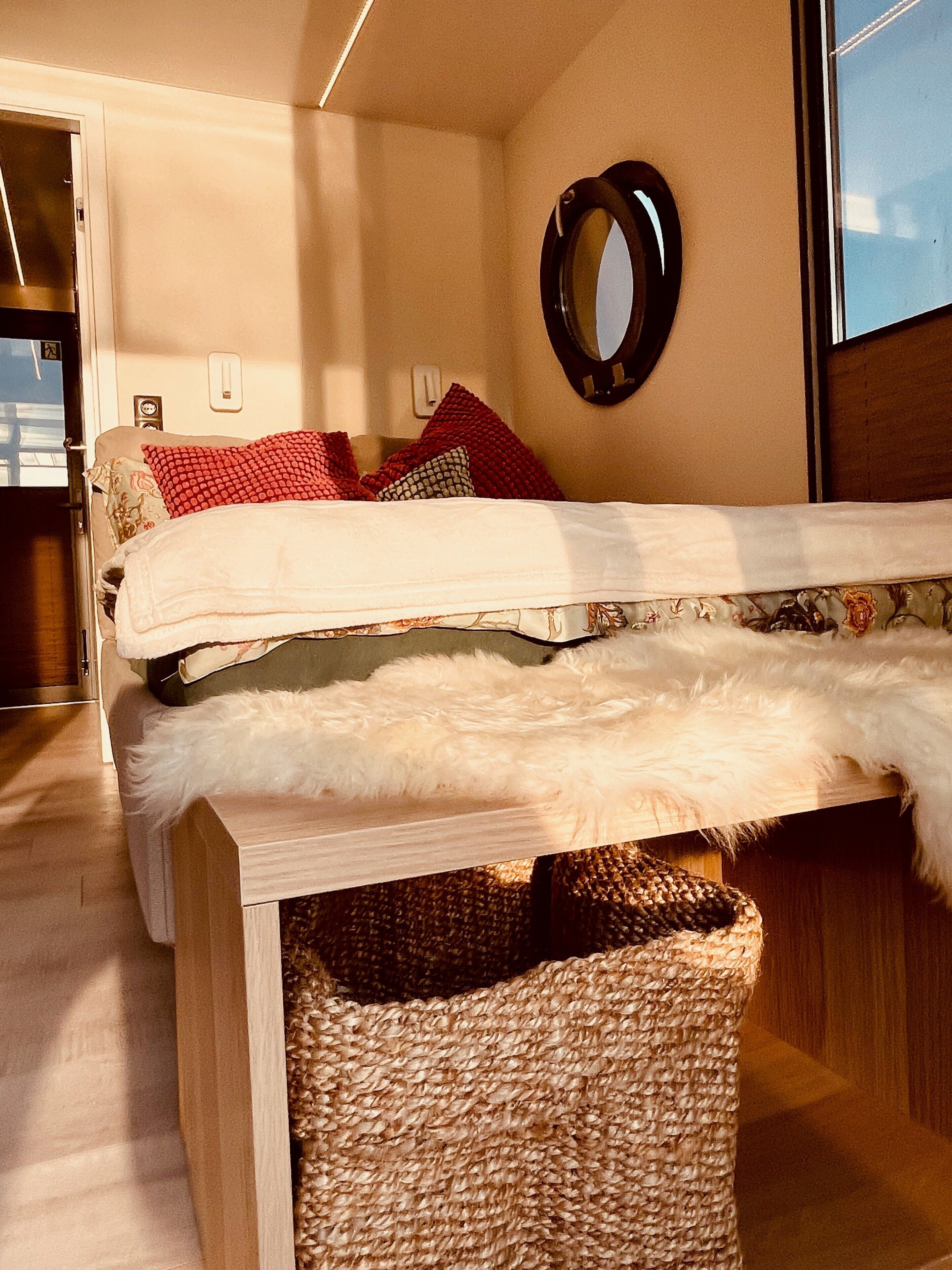 A bed with a white cover, red and patterned pillows, a fluffy white throw, and a woven basket under the bed.