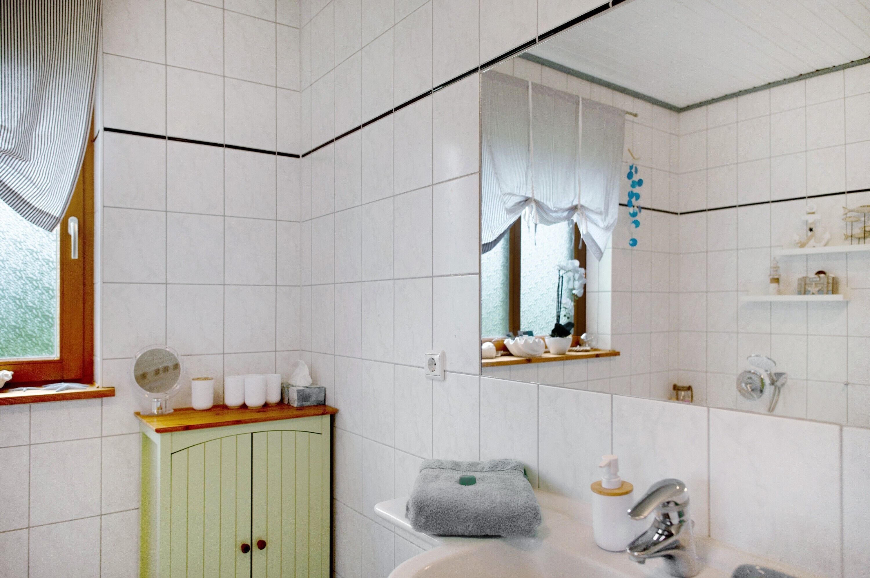 A bathroom with white tiled walls, a green cabinet, a mirror, a sink, and a window with curtains.