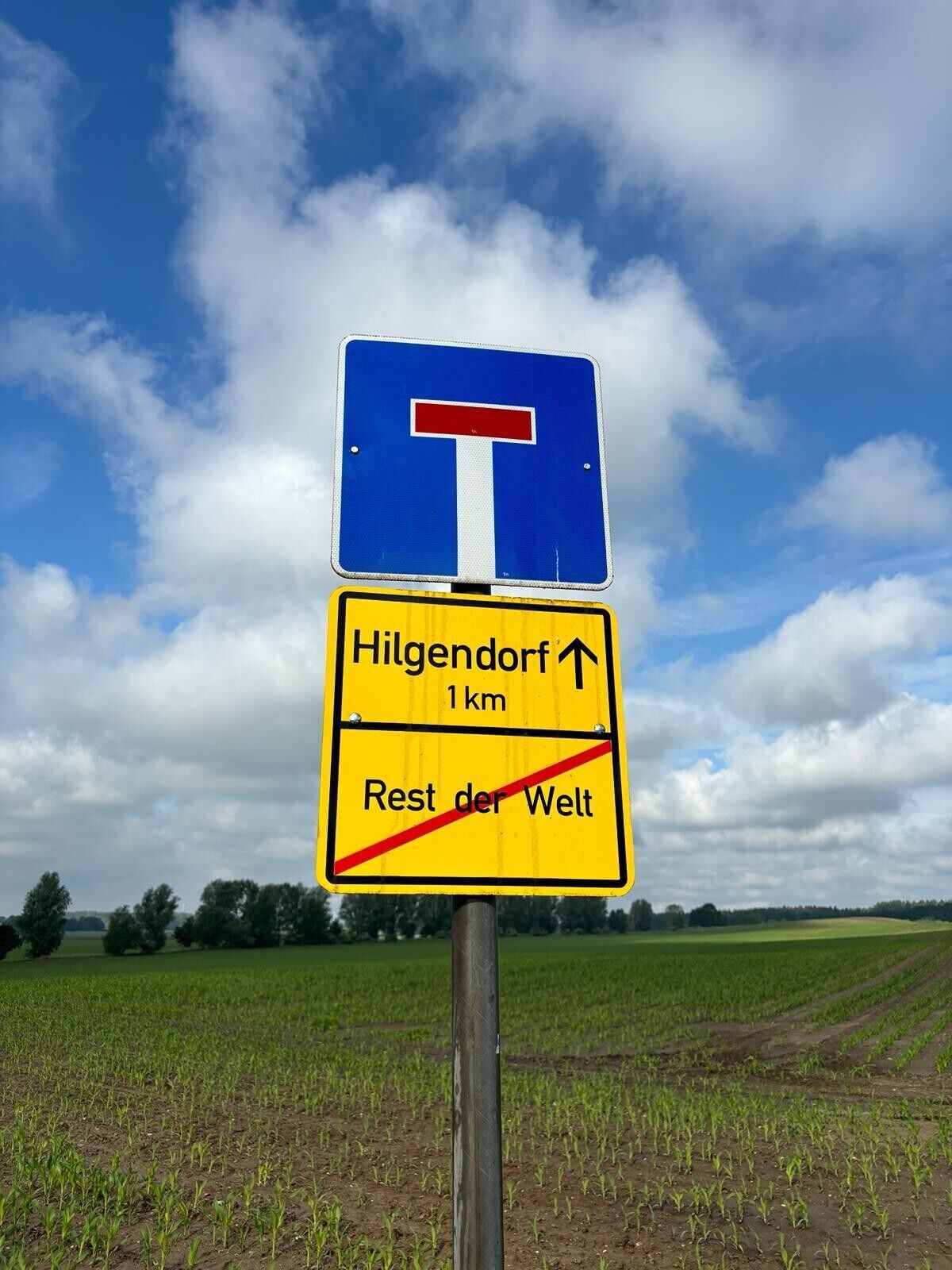 Ein Wegweiser mit blauem Hintergrund und einem roten, T-förmigen Symbol, das eine T-Kreuzung anzeigt. Darunter weist ein gelbes Schild mit schwarzem Text auf Hilgendorf 1 km entfernt und den Rest der Welt, wobei eine rote, diagonale Linie keinen Zutritt anzeigt.