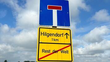 Ein Wegweiser mit blauem Hintergrund und einem roten, T-förmigen Symbol, das eine T-Kreuzung anzeigt. Darunter weist ein gelbes Schild mit schwarzem Text auf Hilgendorf 1 km entfernt und den Rest der Welt, wobei eine rote, diagonale Linie keinen Zutritt anzeigt.