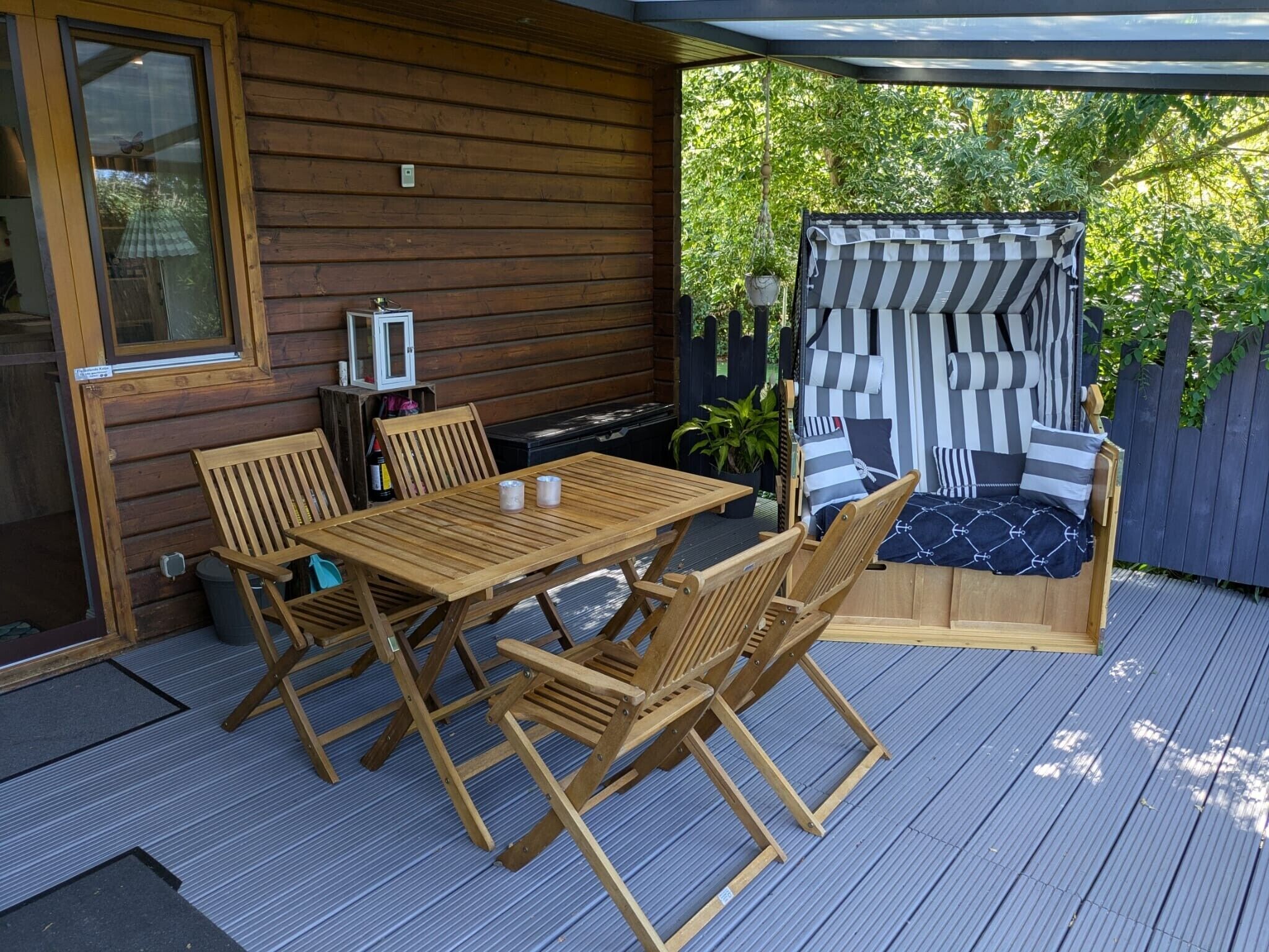 A wooden deck with a table and chairs, a covered seating area with striped cushions, and a view of greenery.