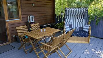 A wooden deck with a table and chairs, a covered seating area with striped cushions, and a view of greenery.
