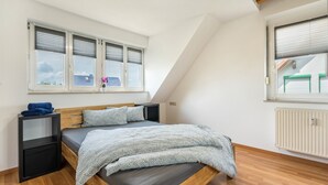 Un dormitorio con cama, suelo de madera, paredes blancas, una ventana con persianas, un radiador y una mesita de noche.