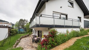 Et toetagers hus med balkon, en anlagt terrasse og en have med blomstrende planter.