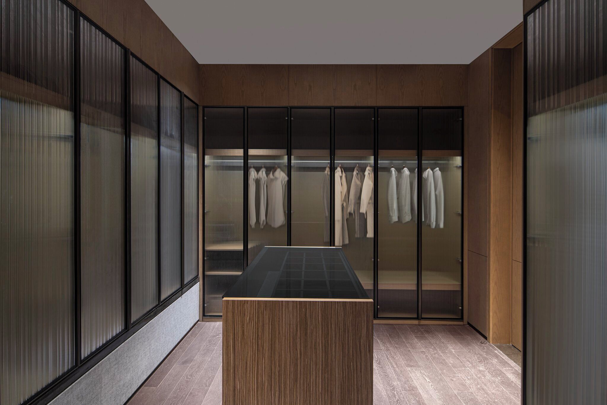 A modern walk-in closet with a wooden bench and a glass door closet containing hanging clothes.