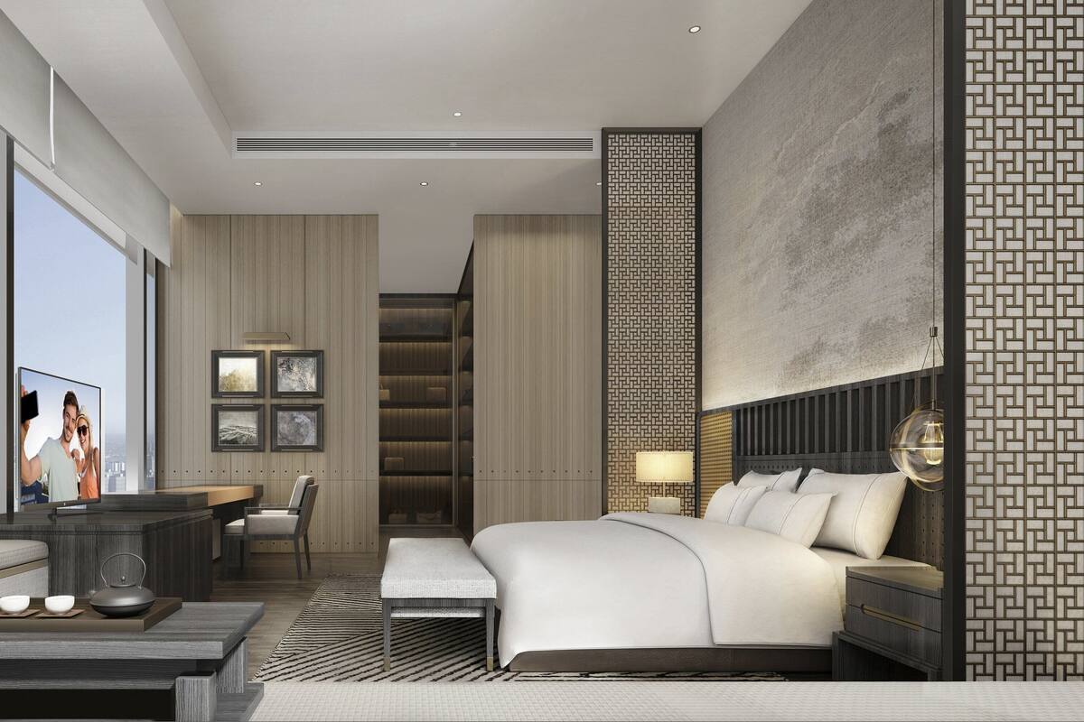 A modern bedroom with a large bed, a desk, and a chair. There is a TV on the desk, and a city view through the window.