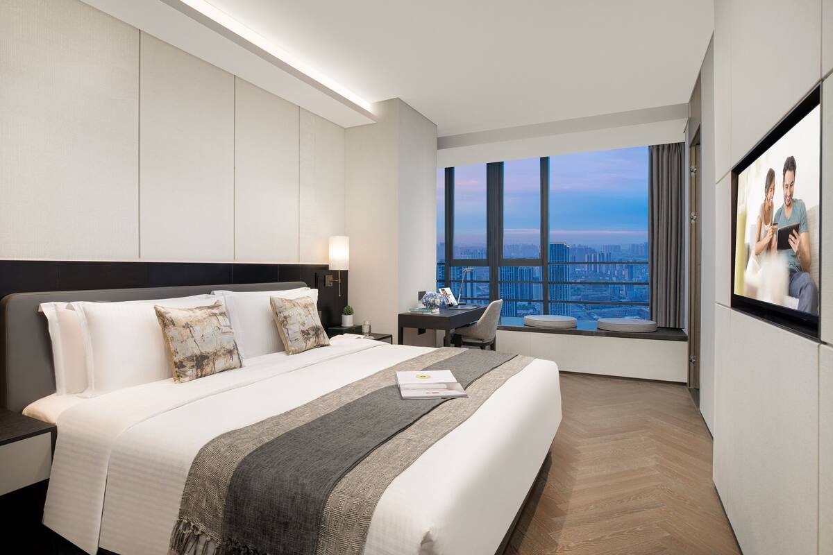 A modern hotel room with a large bed, a desk, and a view of a cityscape through large windows.