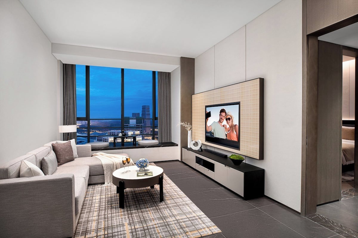 A modern living room with a large TV, a sofa, a coffee table, and a view of the city at dusk.