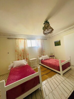 Une chambre avec deux lits, une petite table de chevet, une chaise, une fenêtre avec des rideaux et un luminaire suspendu.