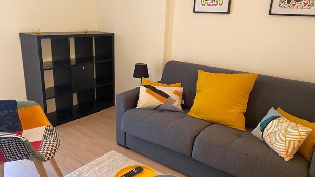 Un salon avec un canapé gris, des coussins jaunes, un meuble noir et une chaise colorée.