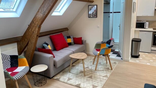 A loft-style living area with a sloped ceiling, a gray sofa with colorful cushions, a small round table, and a colorful chair. A kitchen area with a microwave and a trash can is visible in the background.