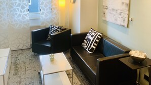 A modern living room with a black leather sofa, a white coffee table, and a patterned throw pillow. A wall-mounted light and a large abstract painting are visible. - Ferienwohnung "ME 7" (Mettmann)