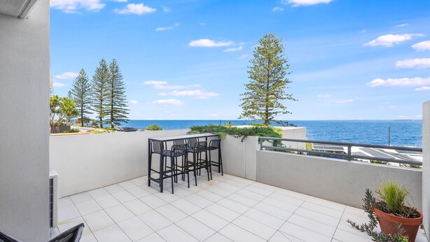 A balcony with a table and chairs, overlooking a coastal view with trees and the sea. - Kings Edge Unit 4 Kings Beach QLD (Kings Beach)